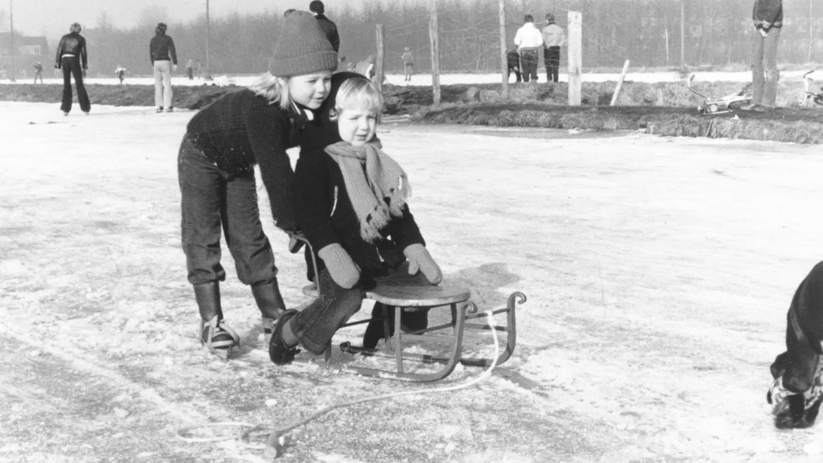 1978 vintage, seventies, retro monochrome portrait of a young girl in duffle coat on Frisian walkers pushing a boy on a retro sled on a natural ice rink.