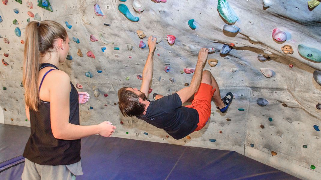 Ein Spotter beim Bouldern schützt die kletternde Person vor Verletzungen, indem er oder sie sicherstellt, dass diese bei einem Sturz auf einem Crashpad landet, am besten mit den Füßen zuerst.