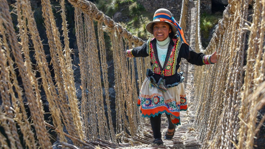 Ein indigenes Mädchen in traditioneller Tracht überquert die Inka-Brücke aus Gras.