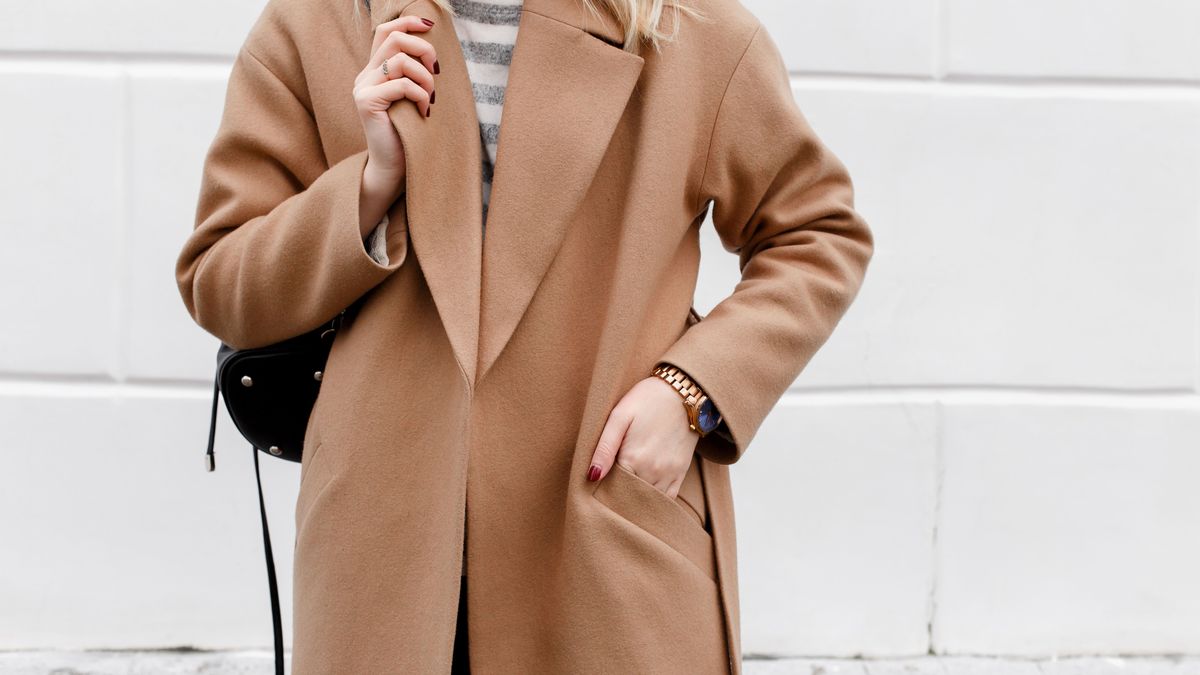 Beautiful young stylish blonde woman wearing beige coat and black backpack posing near white street wall. Trendy casual outfit. Street fashion. Details of everyday look.