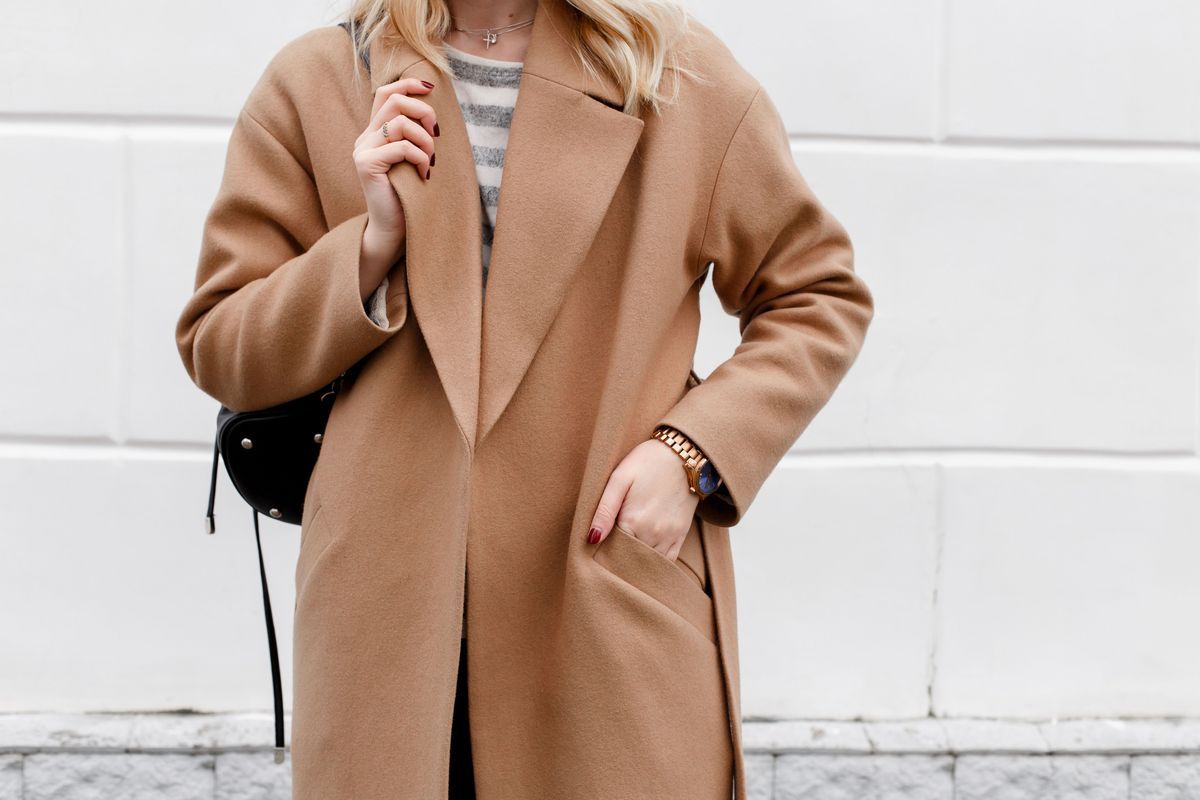 Beautiful young stylish blonde woman wearing beige coat and black backpack posing near white street wall. Trendy casual outfit. Street fashion. Details of everyday look.