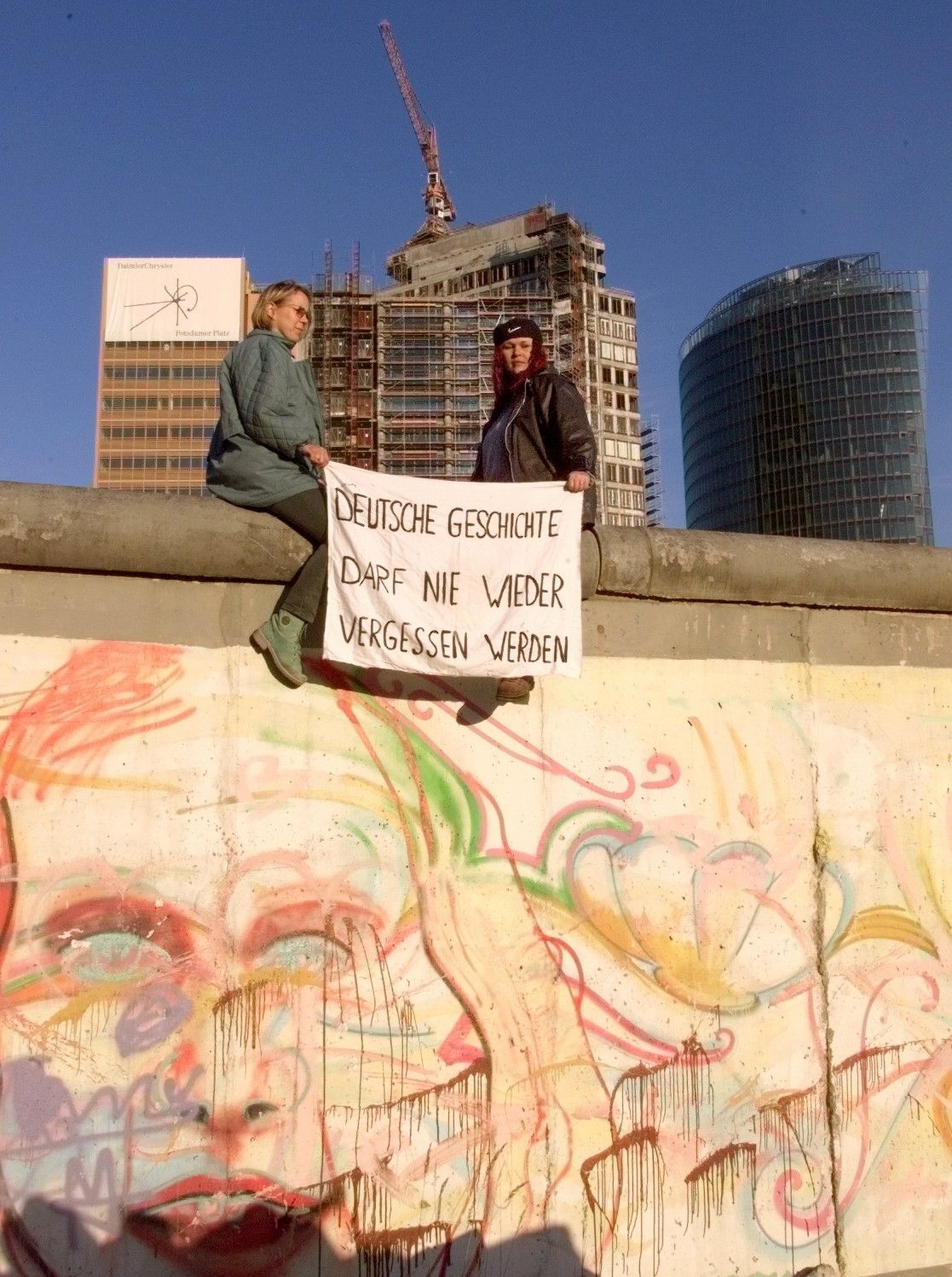 Zwei Demonstrantinnen sitzen am 23. April auf einem der wenigen verbliebenen Teile der Berliner Mauer am Potsdamer Platz in Berlin. Sie verhinderten, dass ein Team einer Abrissfirma damit begann, diesen ursprünglichen Teil der Mauer zu zerstören, um Platz für den Bau einer Büroanlage zu schaffen. Im Hintergrund sind die neu errichteten Firmenzentralen von Daimler und Sony zu sehen.