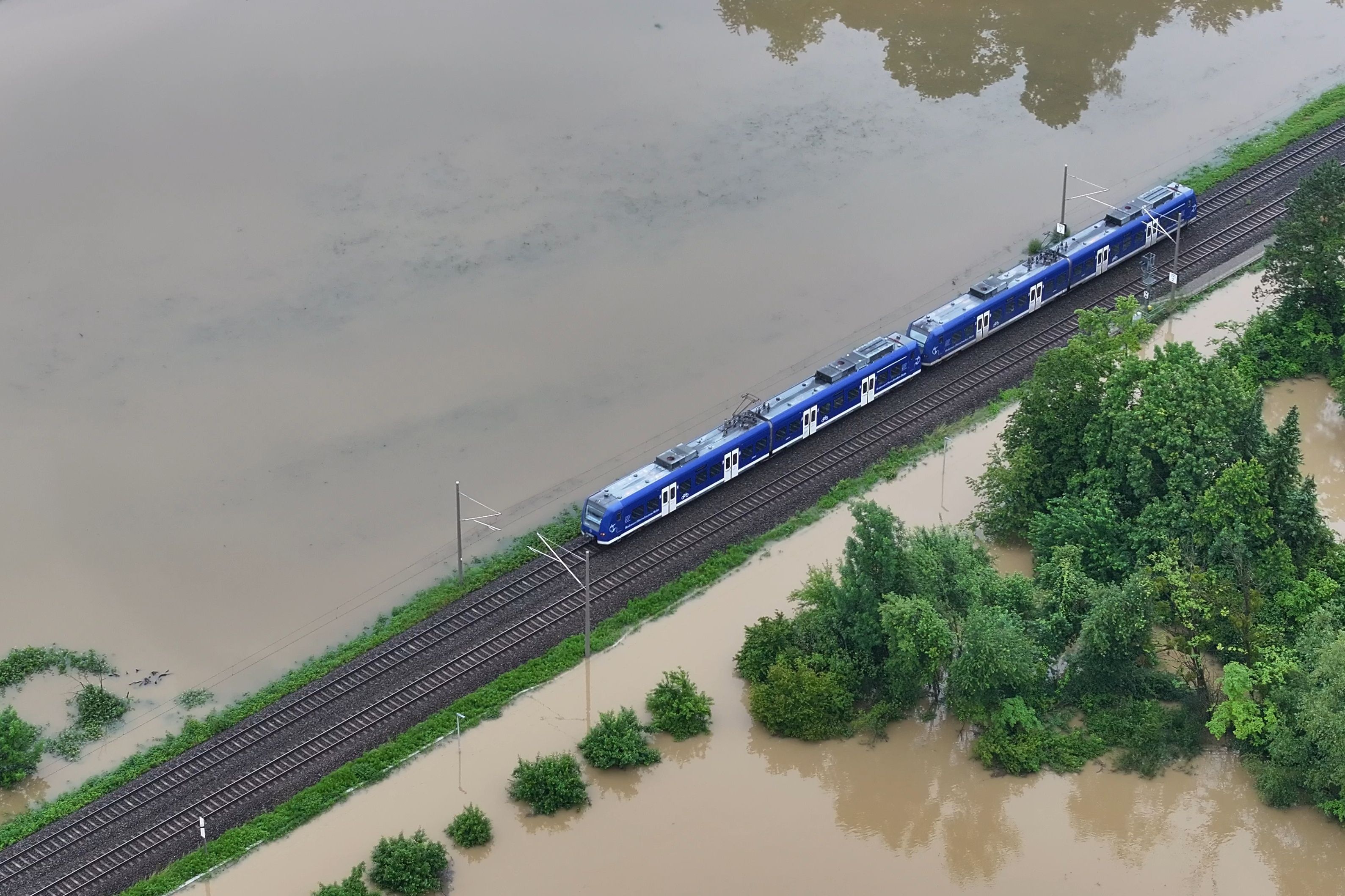Das Hochwasser des Flusses Schussen überschwemmt Teile von Meckenbeuren in Baden-Württemberg. Die Drohnenaufnahme zeigt einen Zug, der über den höher gelegenen Bahndamm fährt.