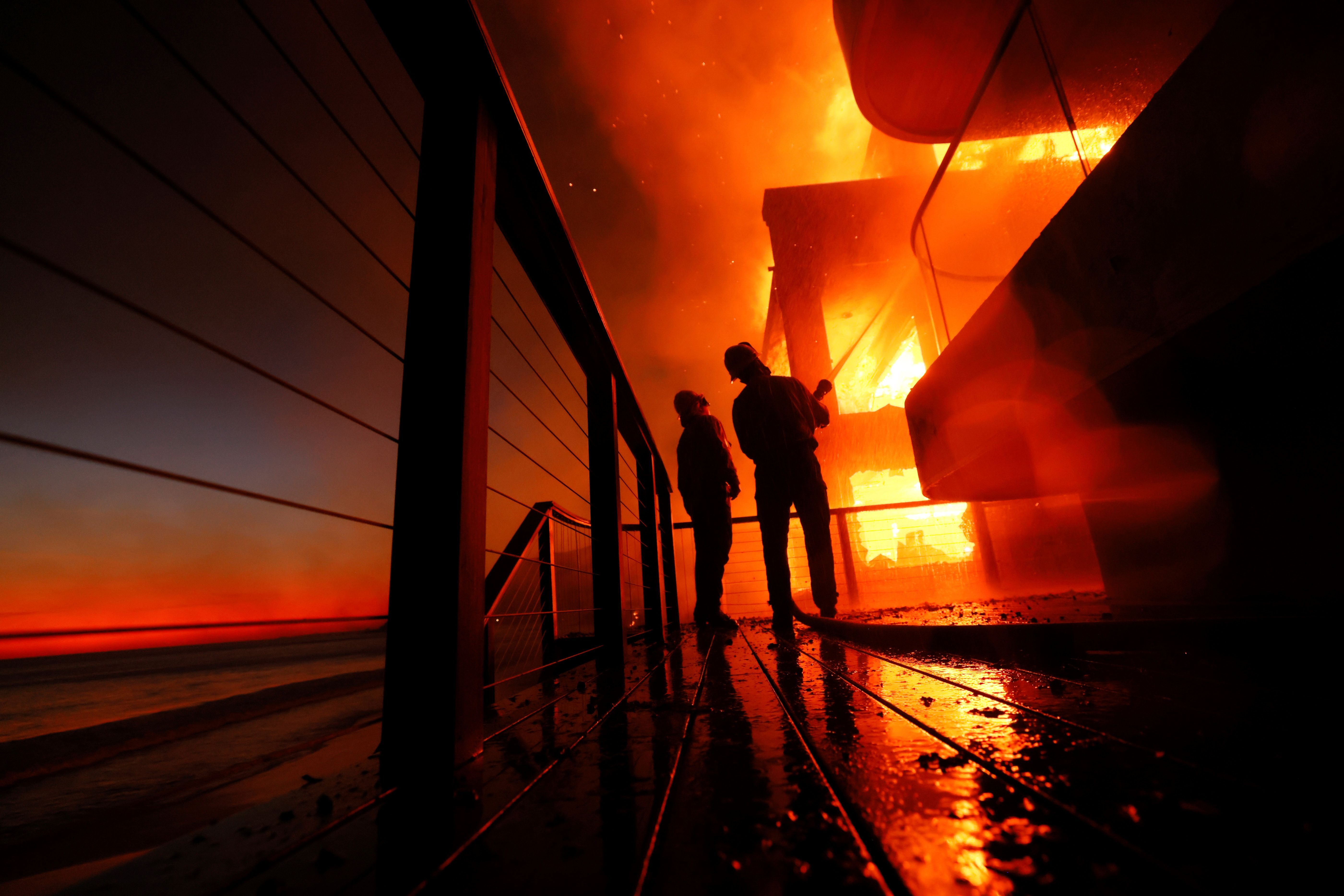 Feuerwehrleute arbeiten von einer Terrasse aus, während das Feuer ein Grundstück am Strand niederbrennt.