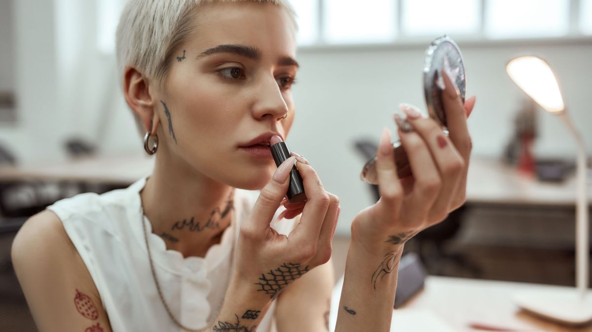 Doing make up. Portrait young and attractive blonde tattooed woman applying lipstick while sitting in the modern office