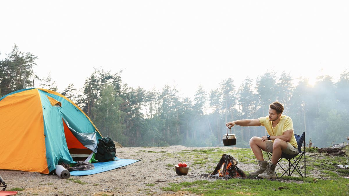 Young man holding kettle over bonfire near camping tent outdoors