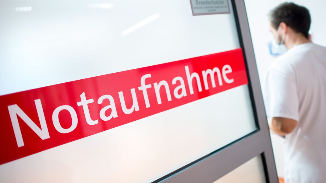 Ein Krankenpfleger steht in einem Krankenhaus neben einer Glastür mit der Aufschrift "Notaufnahme".