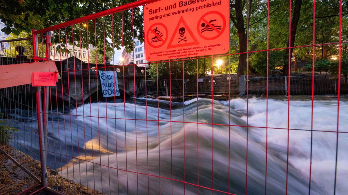 Ein Schild mit der Aufschrift „Surf- und Badeverbot“ hängt an einem Absperrzaun am Ufer der Eisbachwelle im Englischen Garten.