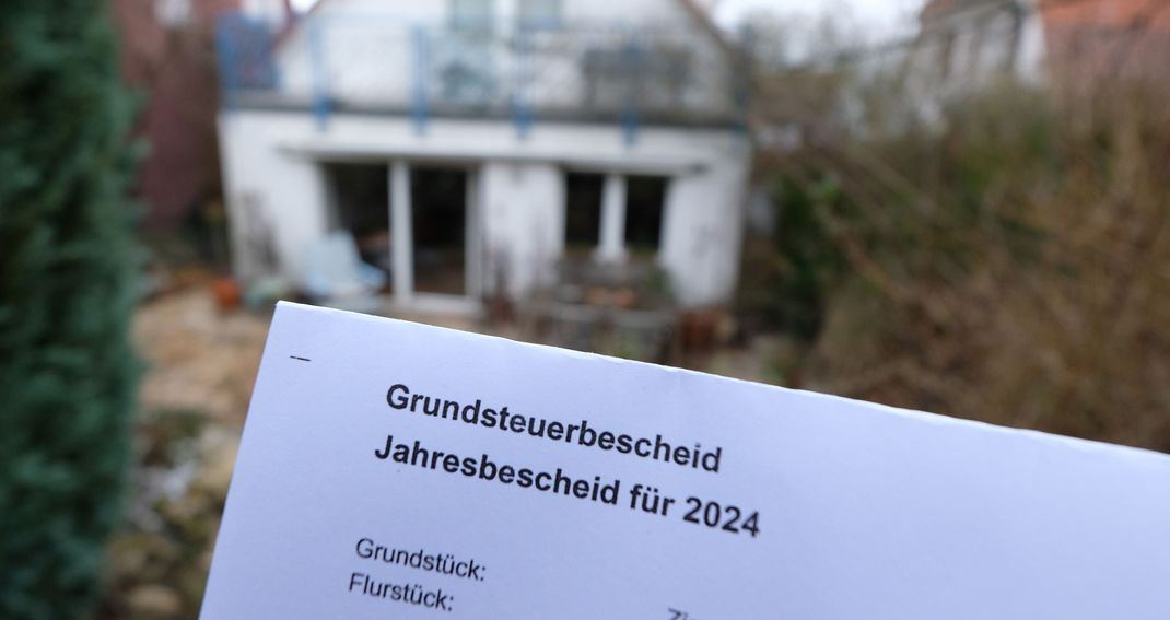 Ein Grundsteuerbescheid für 2024 wird vor einem Privatgrundstück in Stuttgart hochgehalten.