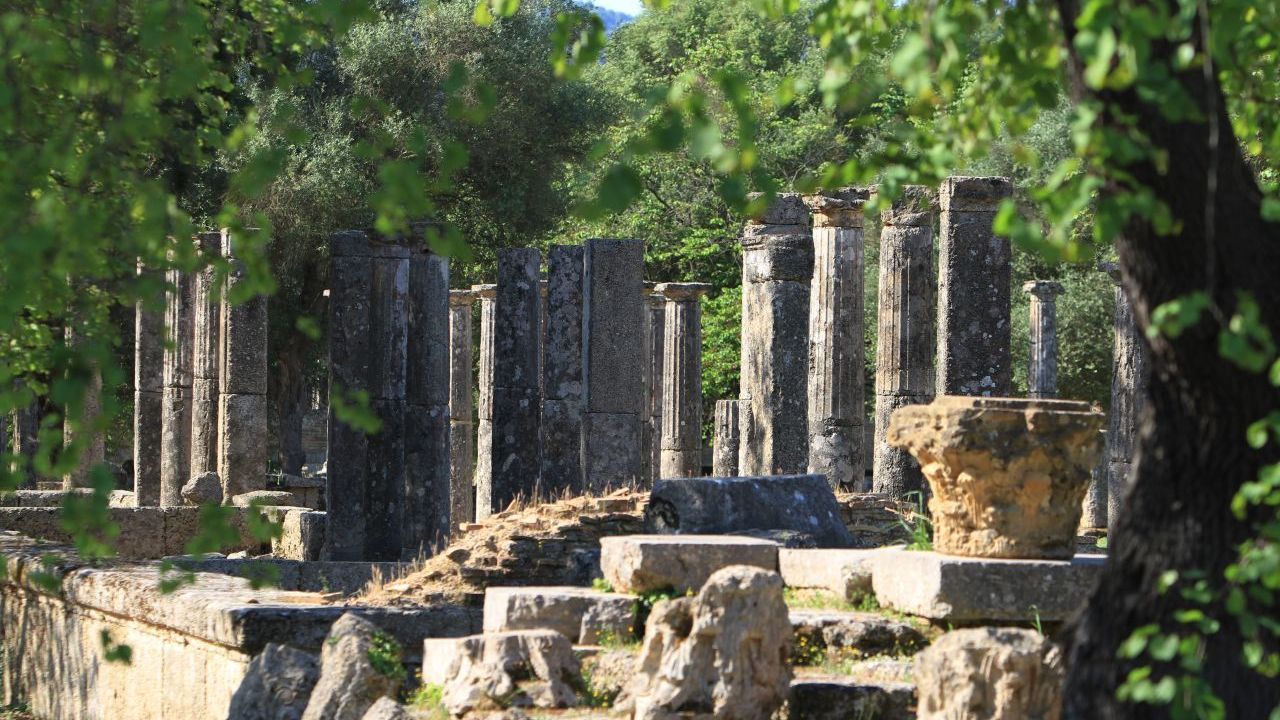 Die Austragungsstätte der Spiele lag in einem Wäldchen, dem heiligen Hain.  Seit 1989 gehört sie zum UNESCO-Weltkulturerbe. Das Foto zeigt im Hintergrund die Säulen der Palaestra. Dies war eine spezielle Trainingsstätte. In der Mitte gab es einen mit Sand bedeckten Innenhof, in dem vor allem die Ringer und Faustkämpfer übten.