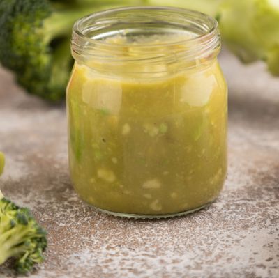 Baby puree with vegetable mix, broccoli, avocado in glass jar on brown concrete, side view, selective focus