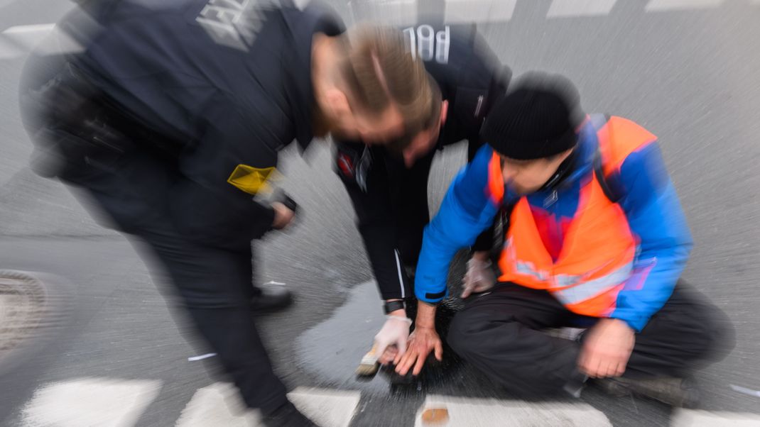 Polizisten versuchen festgeklebte Hände von Aktivisten der Gruppierung Letzte Generation von der Straße abzulösen.