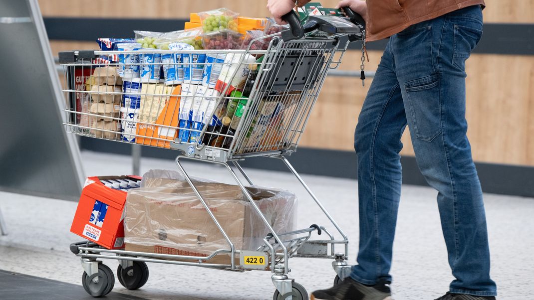 Ein Mann schiebt in einem Supermarkt einen vollen Einkaufswagen mit Lebensmitteln und Getränken.