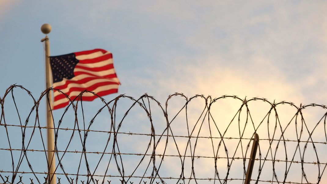 Kuba, Guantanamo Bay: Eine US-amerikanische Flagge weht hinter einem Stacheldrahtzaun im Wind. 