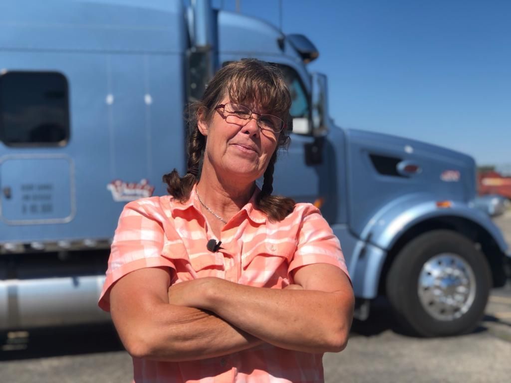 "Trucker Babe" Uschi steht mit verschränkten Armen vor ihrem blauen Truck und lächelt in die Kamera. 